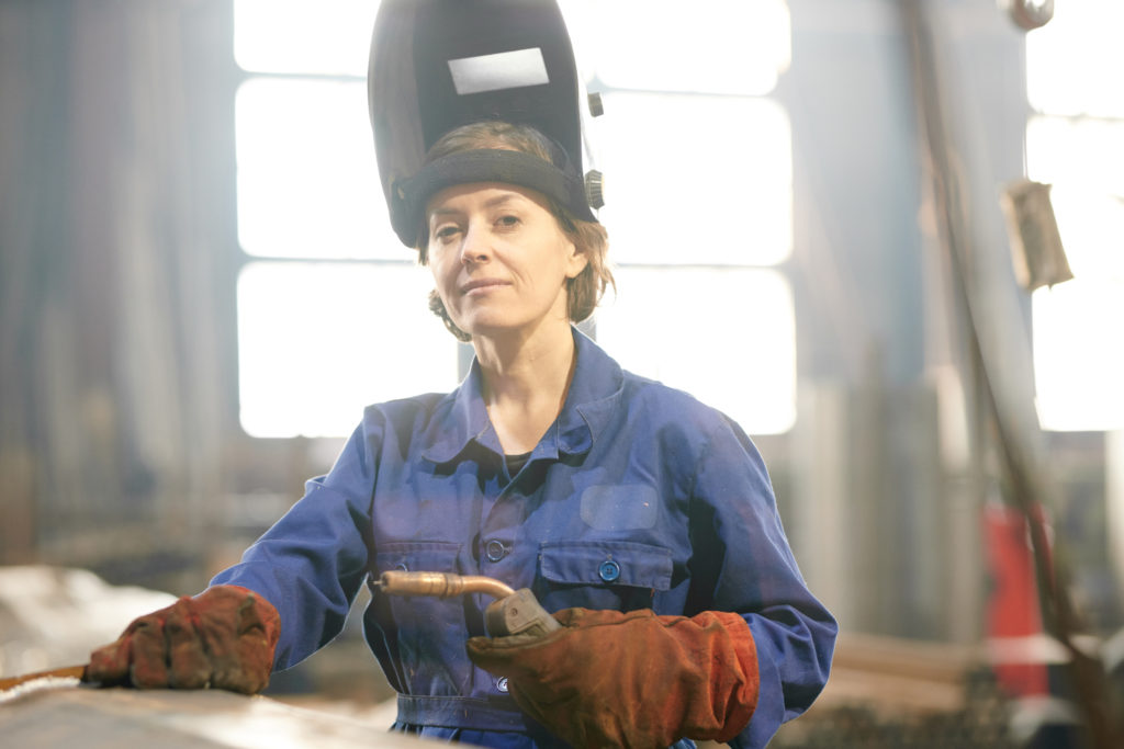 Waist up portrait of smiling female welder posing confidently while working at industrial plant or in garage